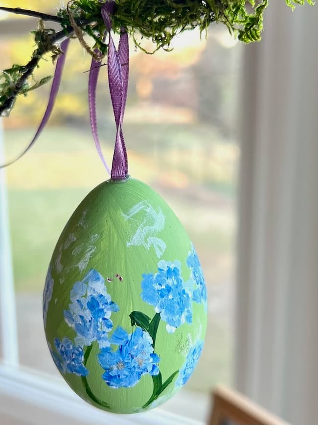 A green egg, adorned with hand-painted blue flowers, hangs from a branch by a purple ribbon. The background reveals a blurred view of a window, offering soft light and hints of spring greenery outside—perfect for any spring decor or Easter crafts project.