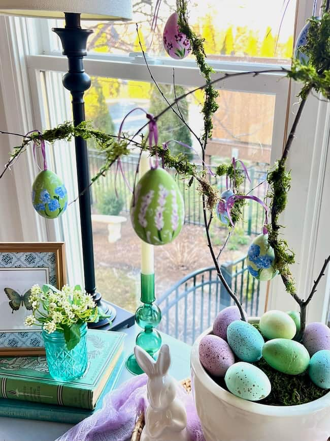 A branch adorned with hand-painted Easter eggs stands in a pot beside a window. A white rabbit figurine nestles next to a bowl of pastel speckled eggs. A stack of books and a green glass with small white flowers complete the cozy spring decor.