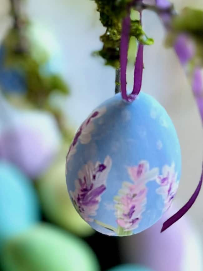 A close-up of a pastel blue Easter egg adorned with hand-painted purple and white flowers. This charming piece of Easter decor hangs elegantly from a purple ribbon, set against a blurred backdrop of green and purple hues, capturing the essence of spring decor.