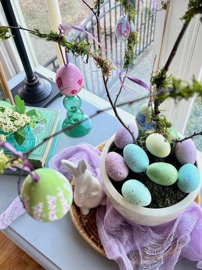 A decorative setup featuring hand-painted Easter eggs in a white bowl on a table. Some eggs are hanging from branches adorned with moss. A ceramic rabbit figurine and books add to the charm of this spring decor, with a window softly illuminating the scene.