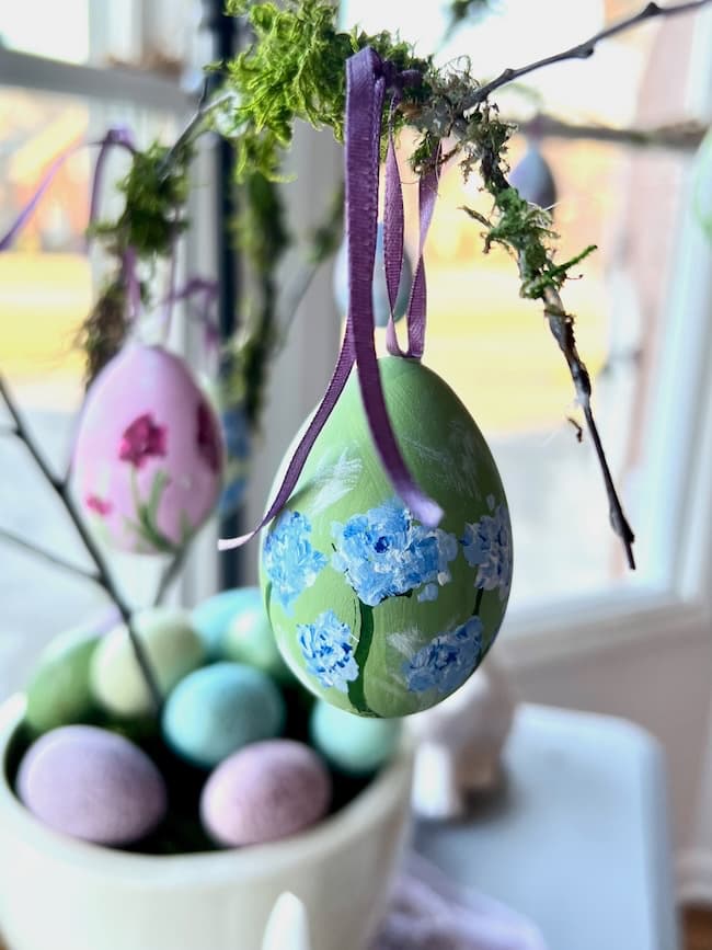 Colorful, hand-painted Easter eggs decorated with floral patterns hang from branches, adorned with purple ribbons. A pot with pastel-colored eggs sits below. Sunlight streams through a nearby window, creating a bright and cheerful spring decor atmosphere.