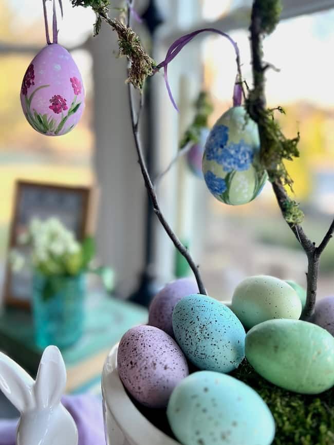 A decorative arrangement exudes charm with hand-painted Easter eggs hanging from a small branch. Below, a variety of colored speckled eggs nestle in a pot, embodying spring decor. In the background, a ceramic rabbit figure and blurred flowers complete the festive scene.