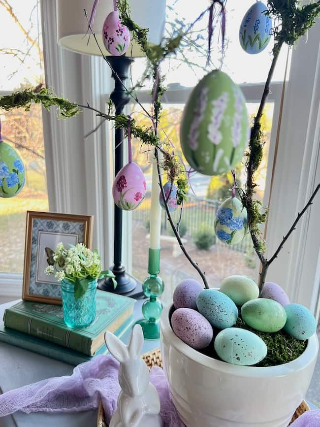 A charming display of Easter decorations features hand-painted Easter eggs hanging from a branch in a white pot brimming with pastel eggs. A white rabbit figurine, green glass vase with flowers, books, and a candle complete the spring decor by the window.