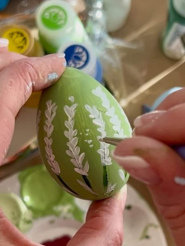 A person meticulously paints a green egg with delicate white floral designs using a fine brush, crafting a beautiful hand-painted Easter egg. Multiple paint bottles and a palette are visible in the background, capturing the essence of spring decor.