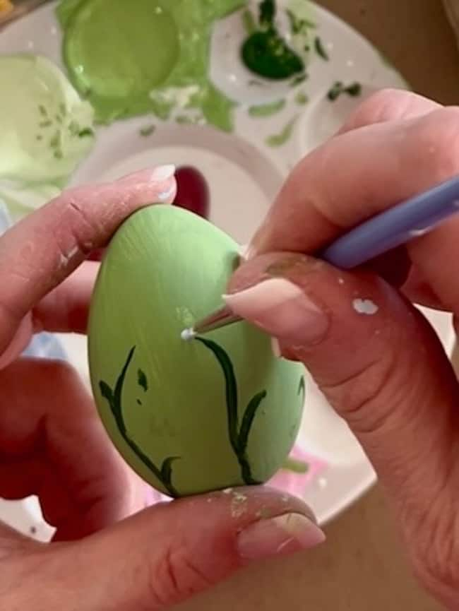 A person is delicately painting a green Easter egg adorned with dark leaf-like lines and a small blue dot using a fine brush. A paint palette filled with various green hues and other colors lies in the background, showcasing the charm of hand-painted Easter eggs perfect for spring decor.