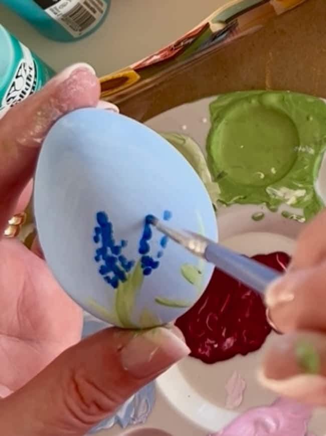 A person is meticulously painting blue flowers with green stems on a light blue egg, crafting exquisite Hand-Painted Easter Eggs. The egg is held in one hand over a palette filled with vibrant spring colors like green and red, perfect for Easter Decor.