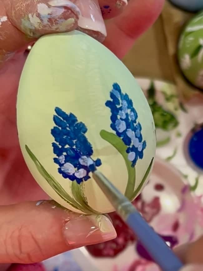 A person holding a painted egg adorned with blue flowers, a hand carefully enhancing its design with a paintbrush. In the background, a vibrant paint palette showcases an array of colors, embodying the essence of hand-painted Easter eggs and charming spring decor.