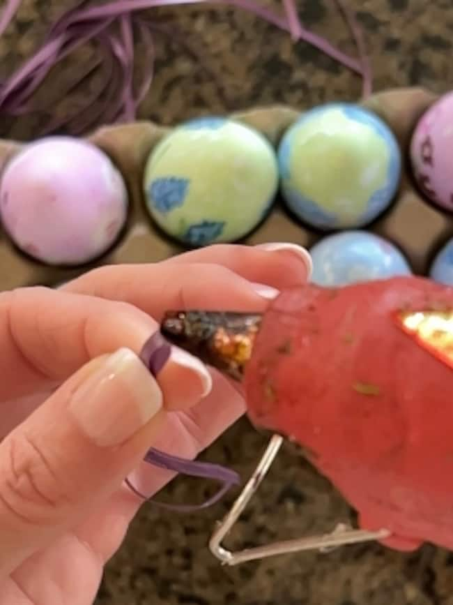 A person holds a purple ribbon near a hot glue gun, crafting delightful Easter decor with hand-painted eggs nestled in the carton behind them.