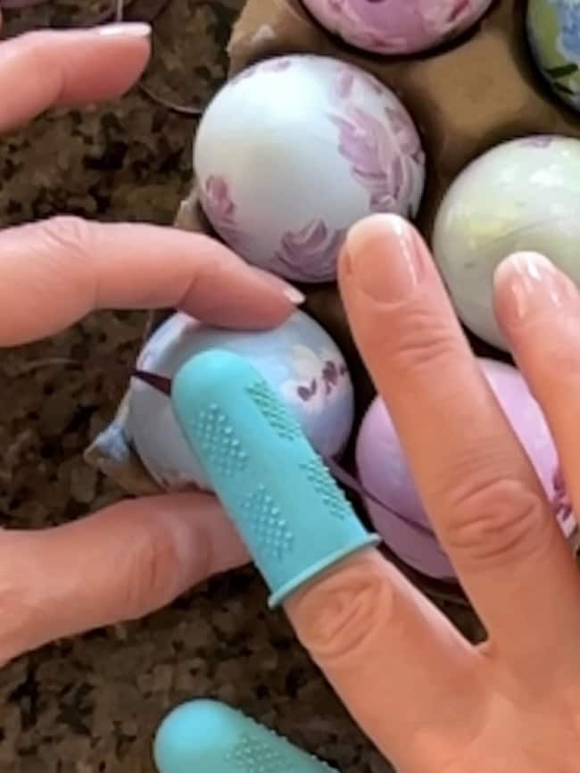 Close-up of hands painting Easter eggs. One hand holds an egg, the other a brush for these hand-painted Easter eggs. The painter sports a blue rubber fingertip cover. Decorated in soft pastel hues, the spring decor eggs rest peacefully in their carton.