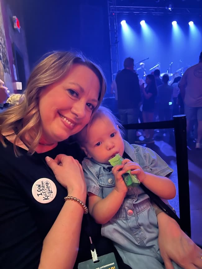 A woman with a badge beams as she holds a child on her lap, the little one savoring a snack. Amidst the buzz of an indoor event, complete with stage lights and eager attendees, they find their own moment of inspiration together.