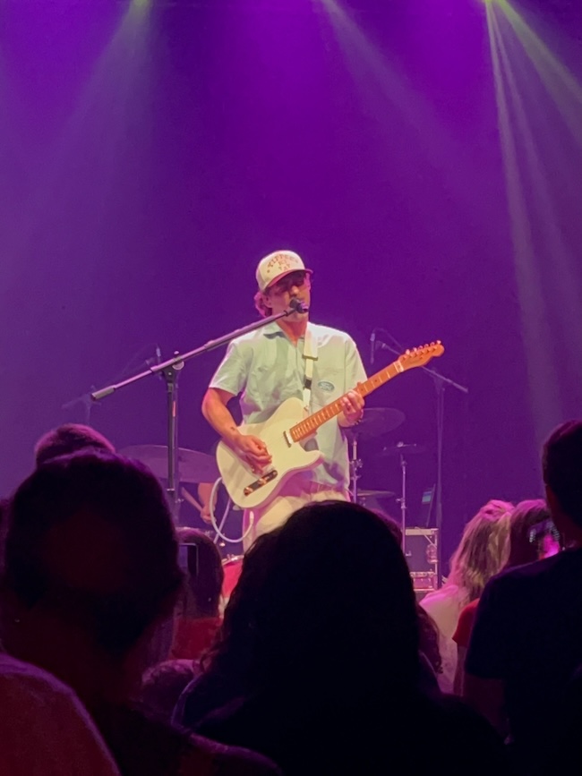 Under the glow of purple lights, a musician captivates the crowd with their electric guitar and soulful voice. Sporting a light blue shirt and baseball cap, they are an inspiration on stage—a true Weekend Find for those seeking musical magic.