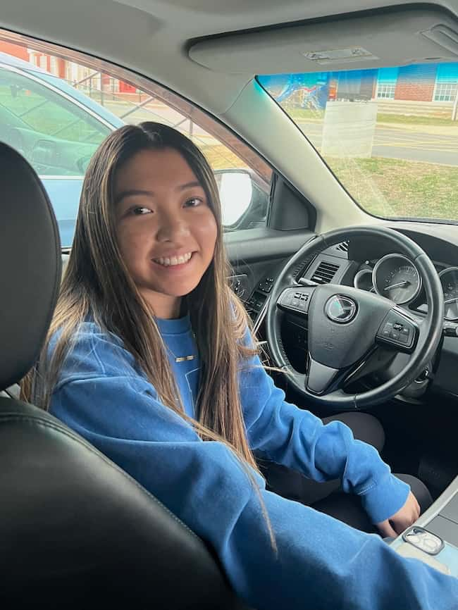 A person with long hair sits in the driver's seat of a car, smiling at the camera, capturing those pretty details. They are wearing a blue sweatshirt. The steering wheel and dashboard controls are visible. The car is parked near a building and another vehicle.