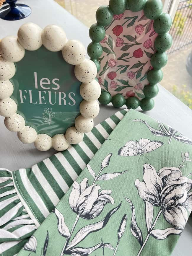 Two decorative frames with green edges: one labeled "les FLEURS," the other adorned with floral designs. Below, a striped green and white fabric and a floral cloth with butterfly motifs rest together, offering weekend inspiration in pretty details on a clean white surface.