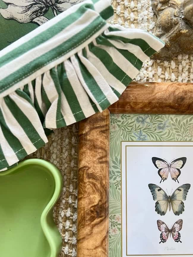 A decorative scene boasts a framed print of three butterflies on a leafy background, complemented by the pretty details of green and white striped fabric with ruffled edges, and a green scalloped-edge dish. The textured woven fabric adds extra charm for some weekend inspiration.