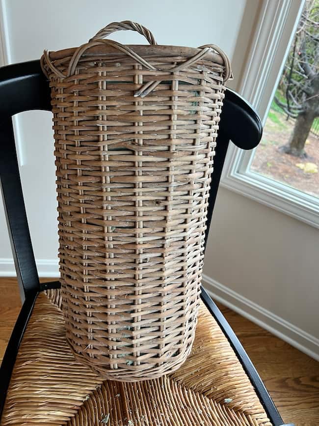 A tall, cylindrical wicker basket, perfect as a spring and summer door flower basket, sits with handles on a woven seat chair near a window overlooking green grass and leafy trees.