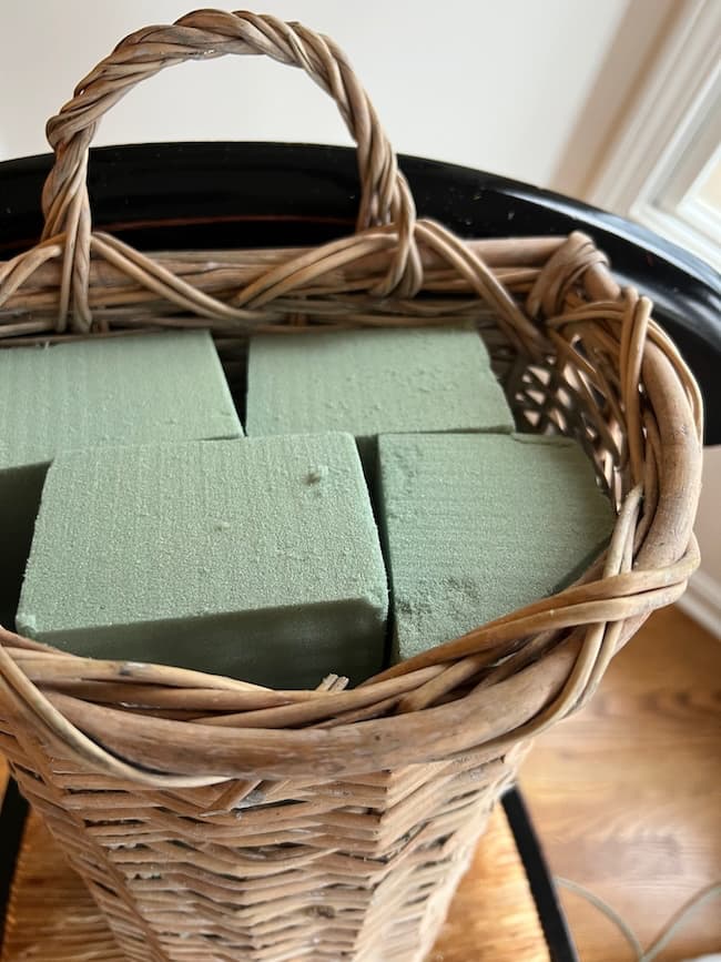 A wicker spring and summer door flower basket with a handle, filled with four green floral foam blocks, sits on a wooden surface near a window.