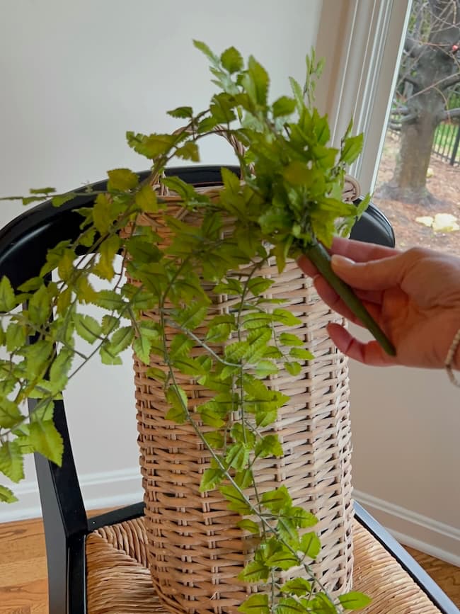 A hand places a green leafy vine into a tall, woven wicker vase—resembling a spring and summer door flower basket—on a black chair near a window.