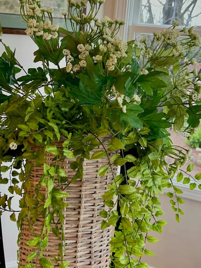 A woven wicker basket, perfect as a spring and summer door flower basket, holds lush green leaves and delicate white flowers, placed indoors near a window with greenery visible outside.