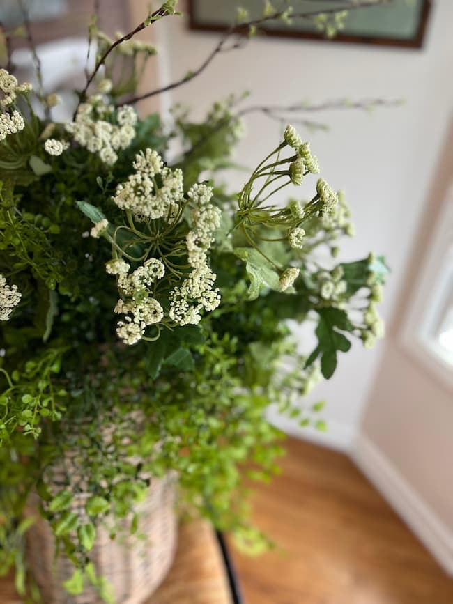 A close-up of a spring and summer door flower basket featuring delicate white flowers and lush green foliage in a woven basket, placed indoors near a window with sunlight streaming in.
