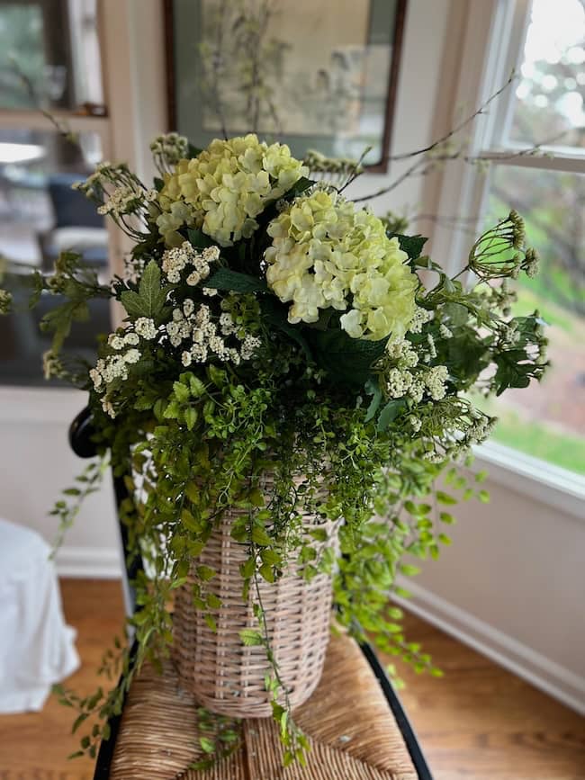 A woven basket vase, inspired by a spring and summer door flower basket, holds lush hydrangeas and greenery, sitting on a wooden chair in a bright, cozy room with large windows.