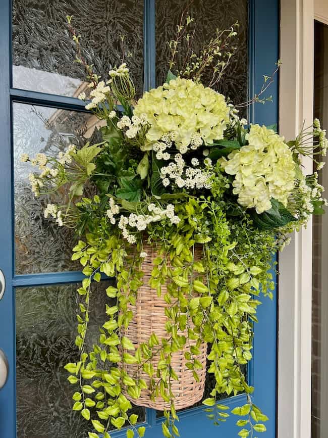 A woven spring and summer door flower basket filled with white hydrangeas, small white flowers, and trailing greenery hangs on a blue front door with glass panels.
