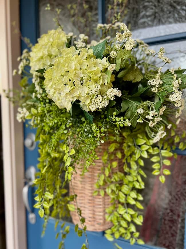 A woven spring and summer door flower basket filled with green and white hydrangeas and leafy greenery hangs on a blue front door with decorative glass panels.