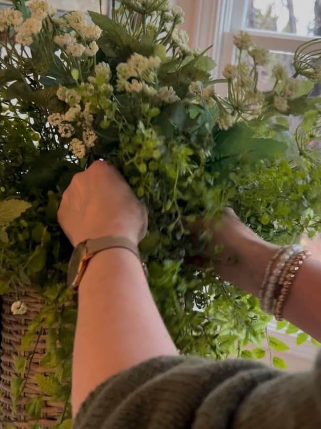 A person wearing a watch and bracelets arranges a lush spring and summer door flower basket filled with green leaves and small white flowers near a sunlit window.