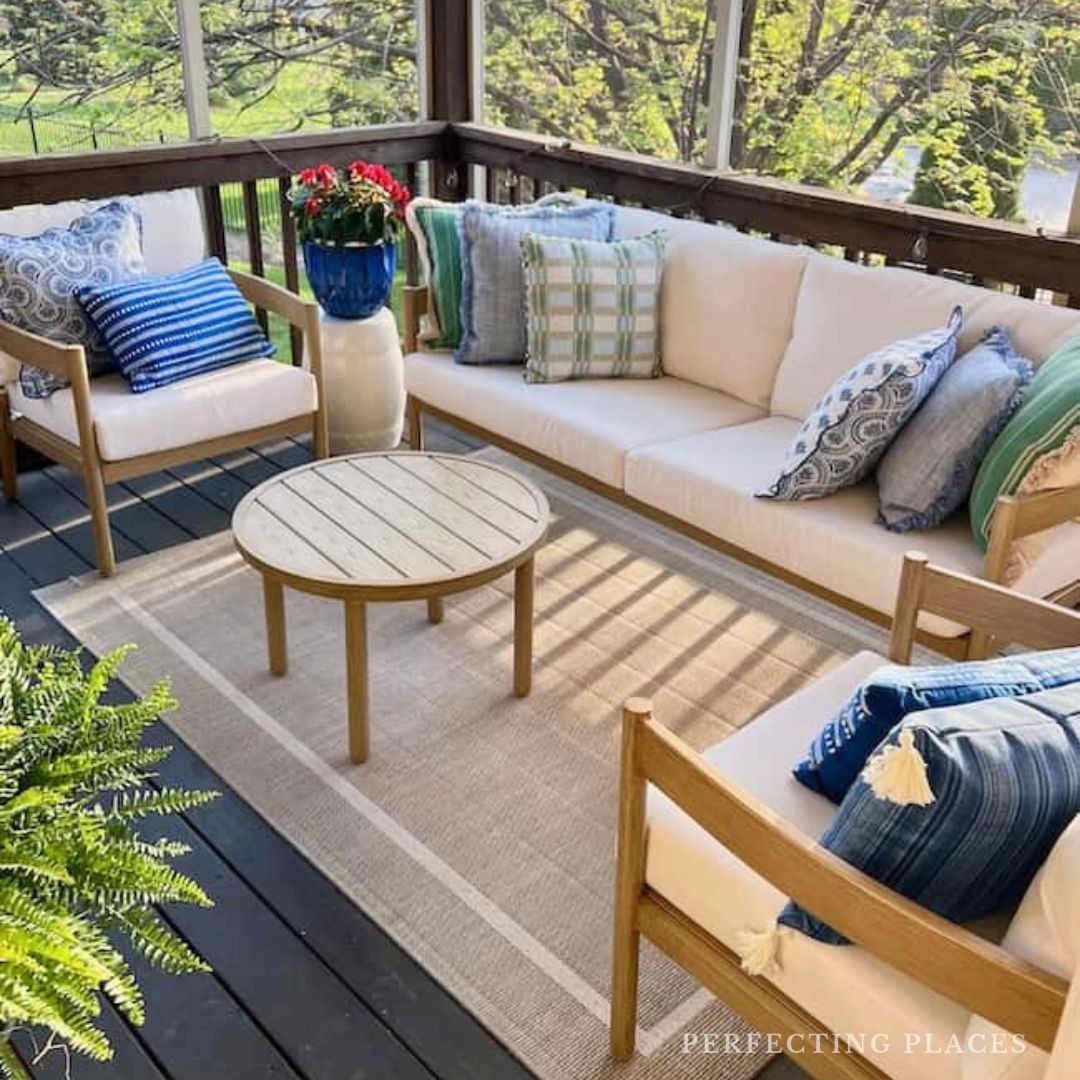 A cozy small porch features cream cushioned seating, blue and green accent pillows, a round wooden coffee table on a beige rug, thoughtful furniture layout, potted plant, and a vase of red flowers—all within a charming screened porch surrounded by trees.