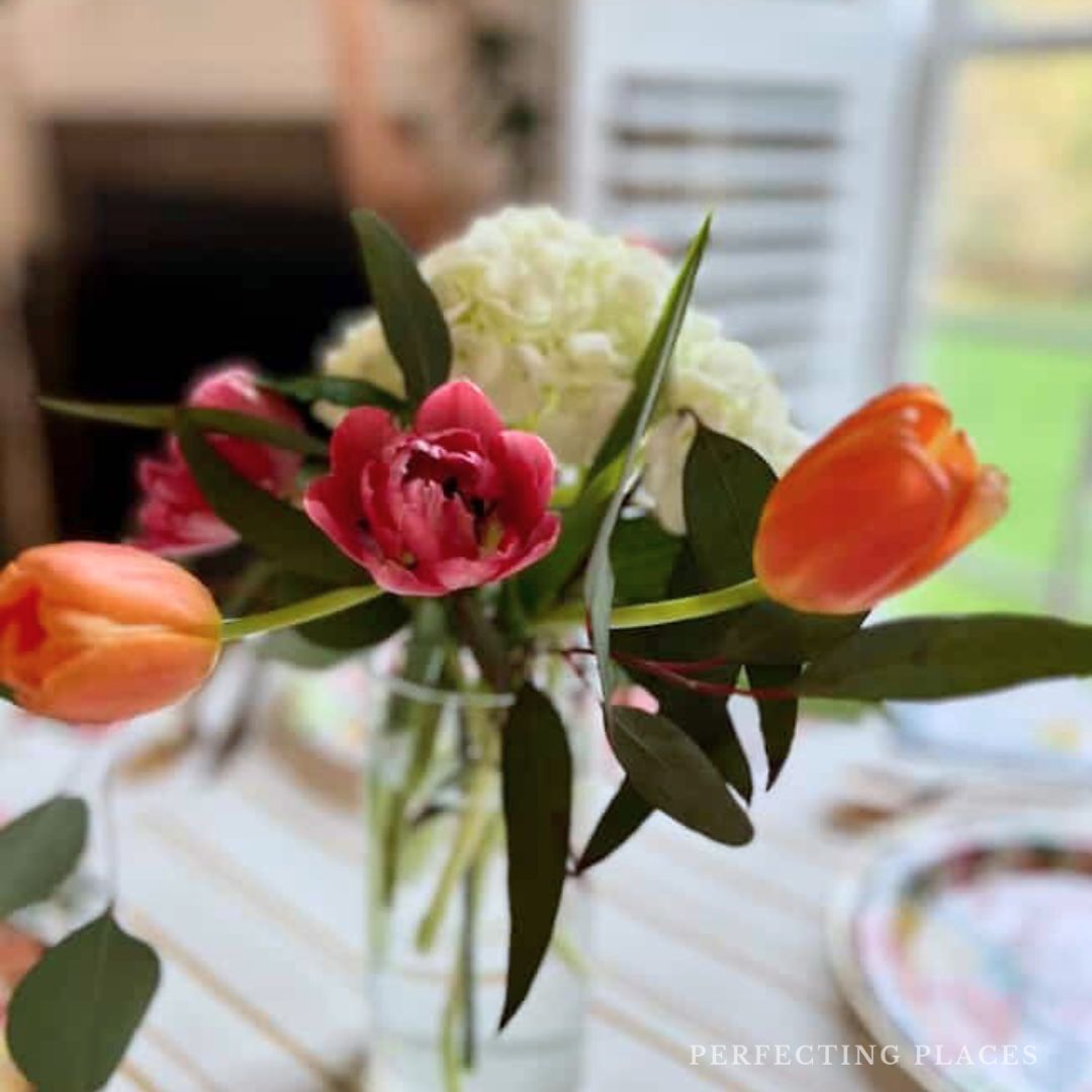 A clear glass vase with orange tulips, a pink peony, white hydrangeas, and green leaves sits on a striped tablecloth near a window with natural light—perfect for April highlights or a Seven on Saturday feature. The background is softly blurred.