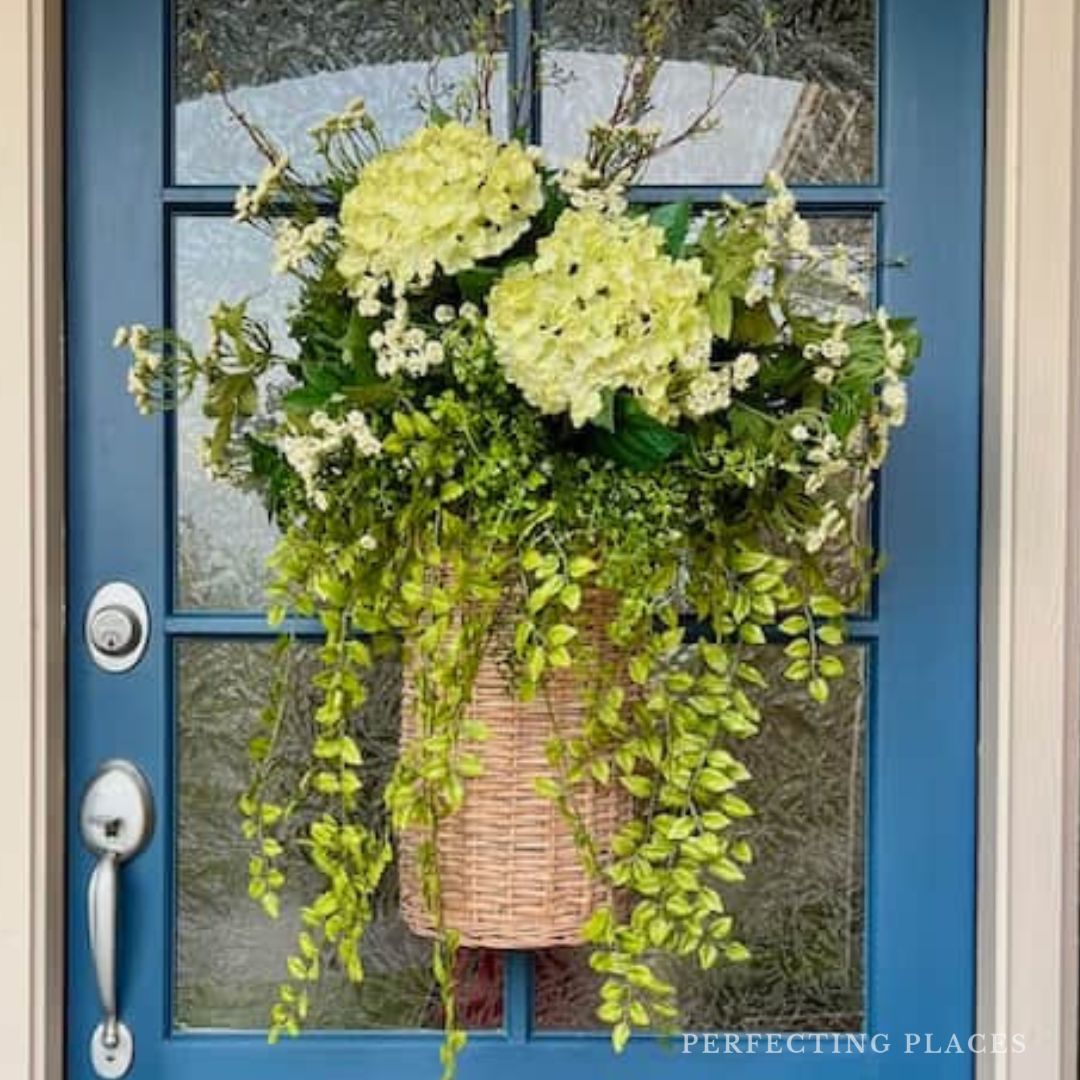 A woven spring and summer door flower basket, filled with green and white blooms and trailing greenery, hangs on a blue front door with glass panels.