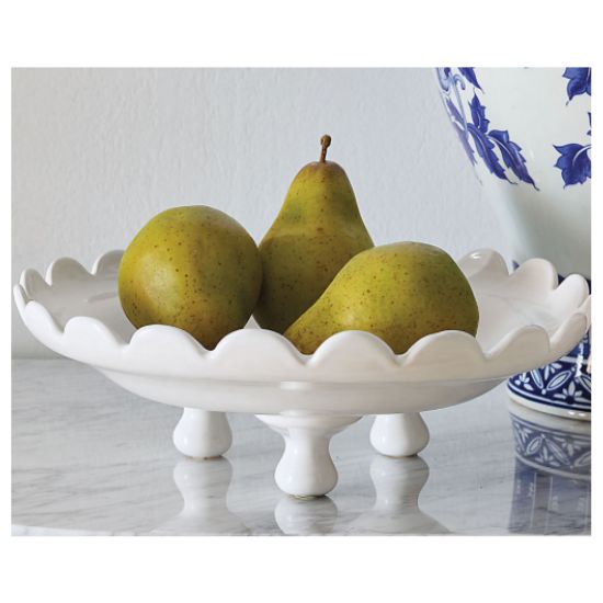 A white scalloped-edge pedestal bowl holding three green pears shows how to decorate a footed bowl, displayed on a marble surface next to a blue and white patterned porcelain vase.