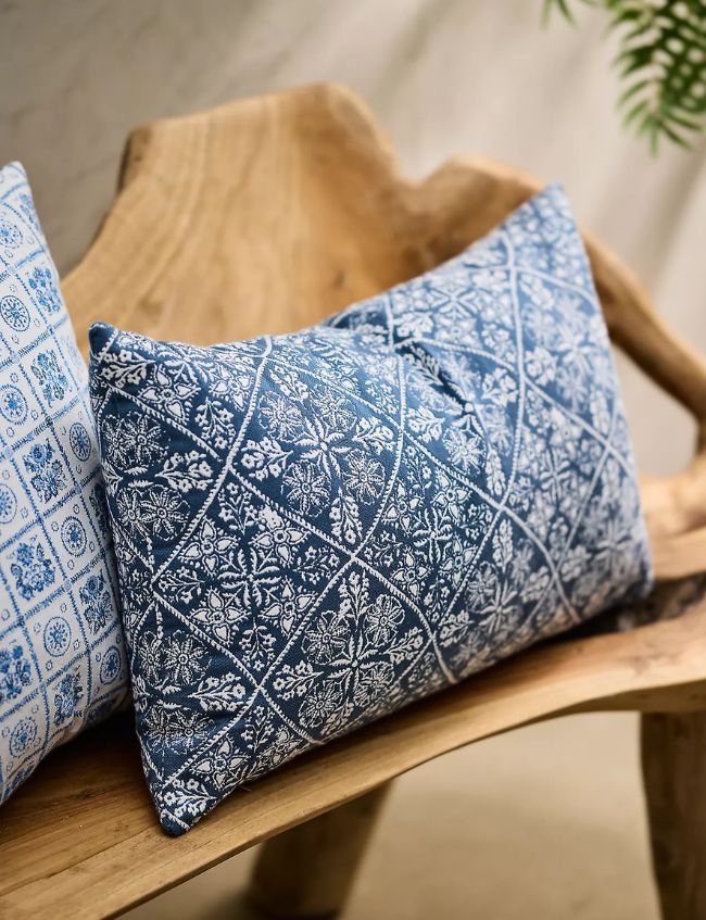 A decorative rectangular pillow with a blue and white floral pattern rests on a rustic wooden bench, next to another pillow with a geometric blue and white design. Discover Top Picks for your home decor—perfect for the Terrain Sale. A green plant is visible in the background.