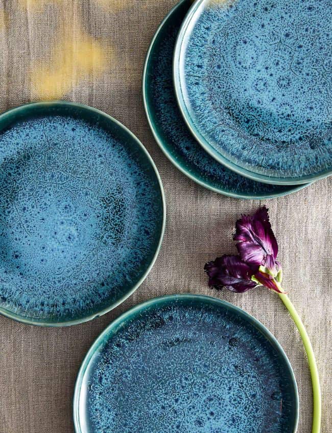 Four blue ceramic plates with speckled patterns are arranged on a beige fabric surface. A single deep purple flower with a green stem lies near the bottom right plate—one of our Top Picks for the Terrain Sale.