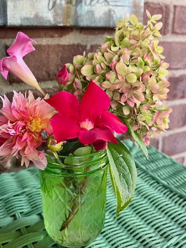 A small green glass jar holds a bouquet of flowers, including a bright red bloom, a pink trumpet-shaped flower, a pink dahlia, and a cluster of green and pink hydrangea, set on a green woven surface against a brick wall.