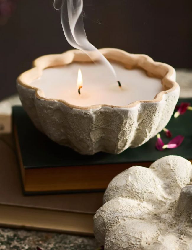 A stone-textured bowl-shaped candle with two burning wicks sits atop a closed book, as wisps of smoke rise from one wick. Pink flower petals and another stone object are nearby—an elegant addition to your Top Picks this season.