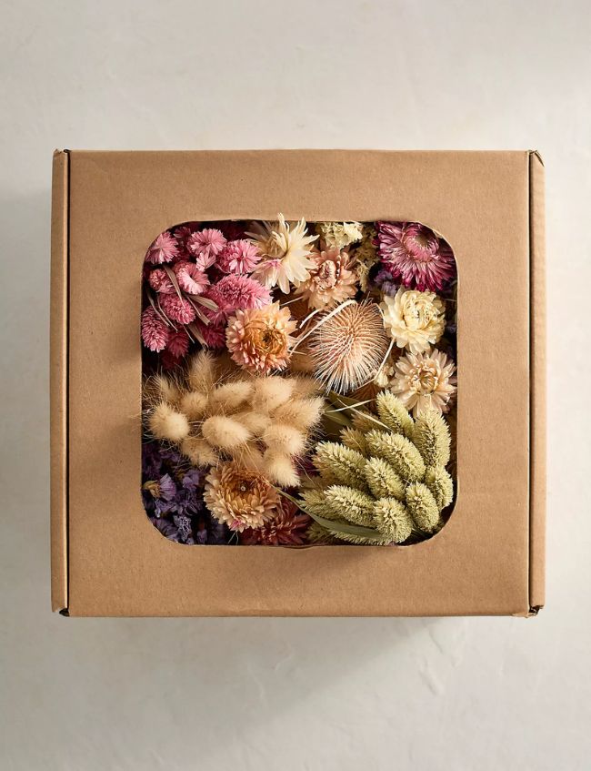 A brown cardboard box with a clear window on top reveals an assortment of colorful dried flowers—pink, purple, cream, and green varieties—arranged neatly inside. Discover this item among our Top Picks in the Terrain Sale.