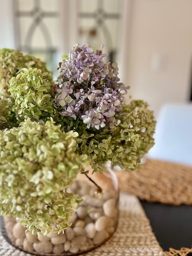 A glass vase filled with round stones holds green and purple hydrangea flowers, sitting on a woven placemat on a table in a softly lit room.