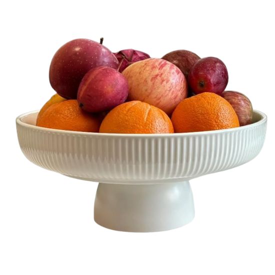 A white pedestal fruit bowl, perfect for showcasing how to decorate a footed bowl, is filled with apples, oranges, and plums against a plain white background.