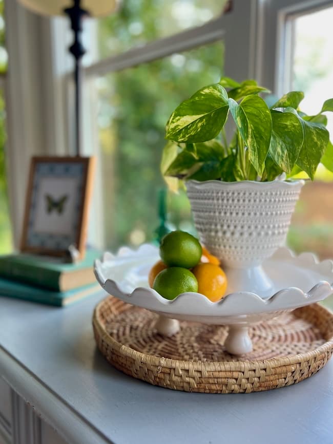 A white planter with a green leafy plant sits on a scalloped cake stand, offering inspiration for how to decorate a footed bowl, next to limes and an orange on a woven tray atop a white windowsill. Blurred books and a butterfly frame complete the scene.