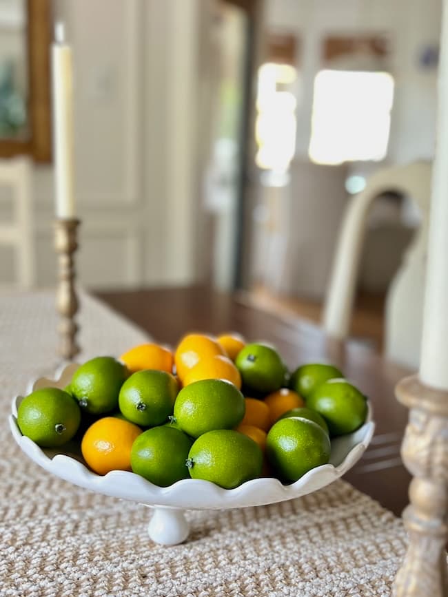 A white scalloped bowl, styled as an example of how to decorate a footed bowl, is filled with green limes and yellow lemons on a woven table runner, flanked by tall candlesticks and a softly blurred dining room in the background.