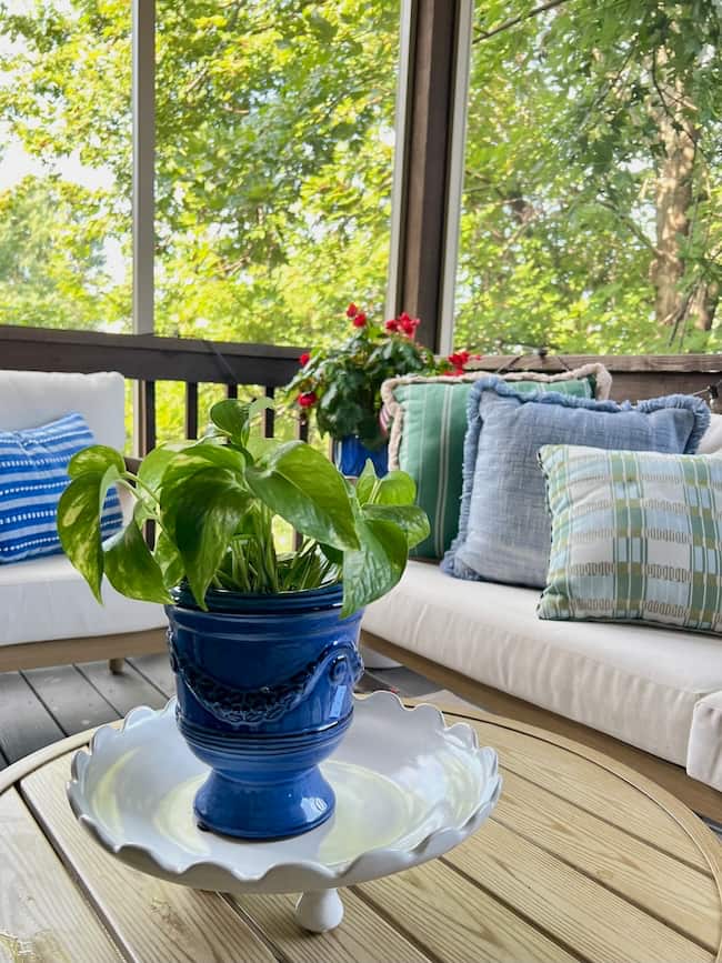 A leafy green potted plant in a blue ceramic pot sits on a white decorative tray atop a wooden table, inspiring ideas on how to decorate a footed bowl, with cushioned outdoor sofas and green trees in the background.