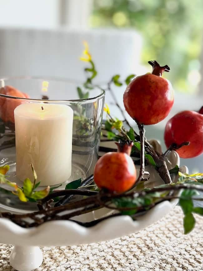 A white candle in a glass holder sits on a white pedestal tray, surrounded by artificial pomegranate branches and green leaves—an inspiring example of how to decorate a footed bowl with elegance against a softly blurred background.