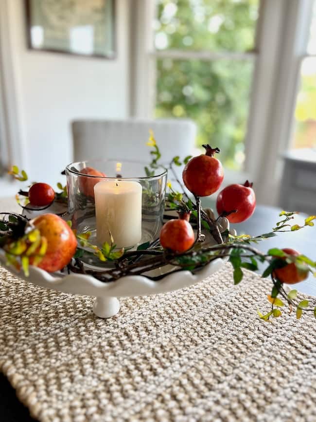 A white pedestal tray is styled like how to decorate a footed bowl, with a glass candle holder and lit white candle, surrounded by artificial red pomegranates and green foliage on a textured beige placemat.