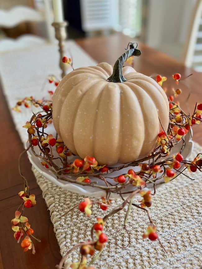 A light beige pumpkin sits on a white plate, surrounded by a ring of orange and yellow faux berries—an inspiring idea for how to decorate a footed bowl—atop a woven table runner on a wooden dining table.