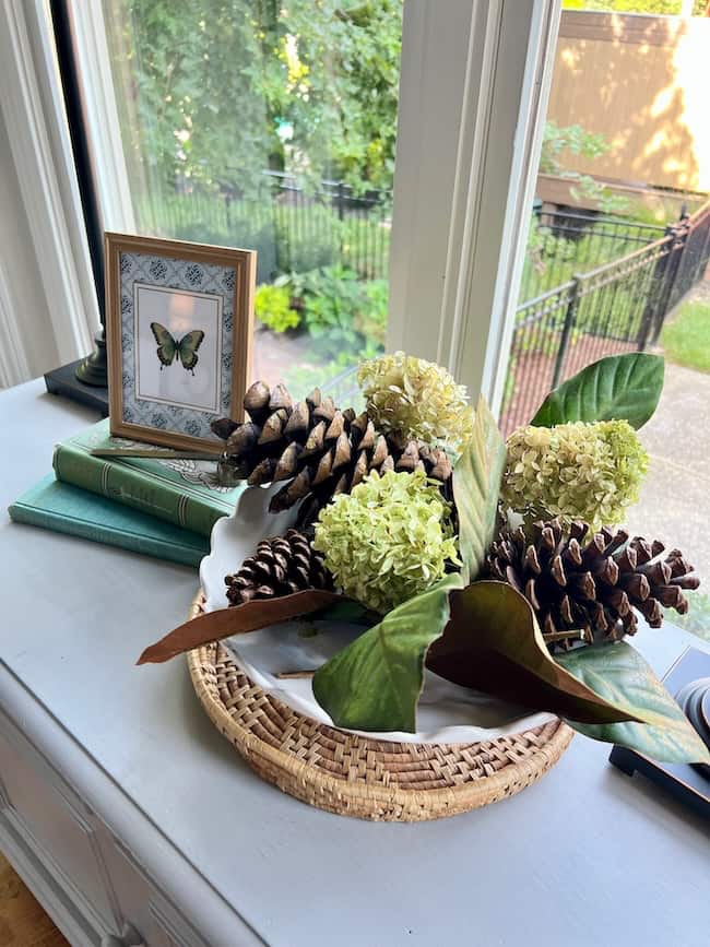 A woven tray displays pinecones, white hydrangea flowers, and green leaves—offering inspiration on how to decorate a footed bowl—while stacked books and a framed butterfly illustration rest nearby. A garden view completes the scene.