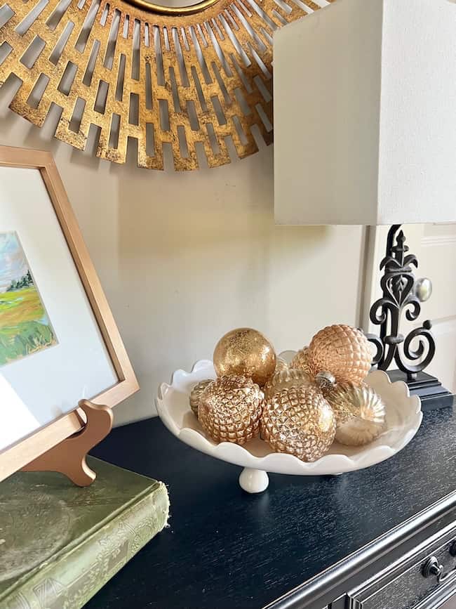 A white footed bowl filled with gold and rose gold textured ornaments sits on a black table beside a green book, a framed painting, a lamp with a white shade, and a gold sunburst mirror—showcasing how to decorate a footed bowl elegantly.