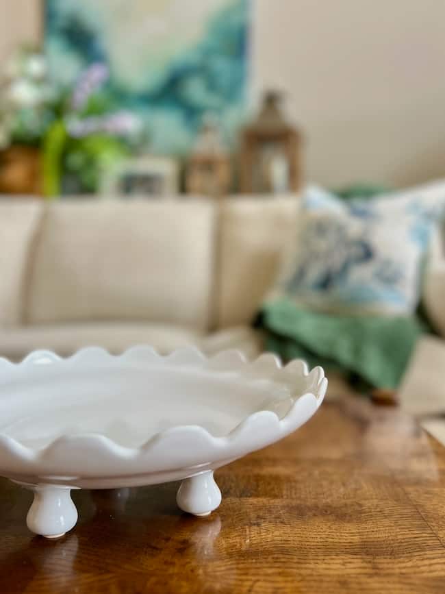 A white, wavy-edged ceramic footed bowl sits on three short legs atop a wooden table in front of a beige couch with blue and green accent pillows—an inviting scene for inspiration on how to decorate a footed bowl.