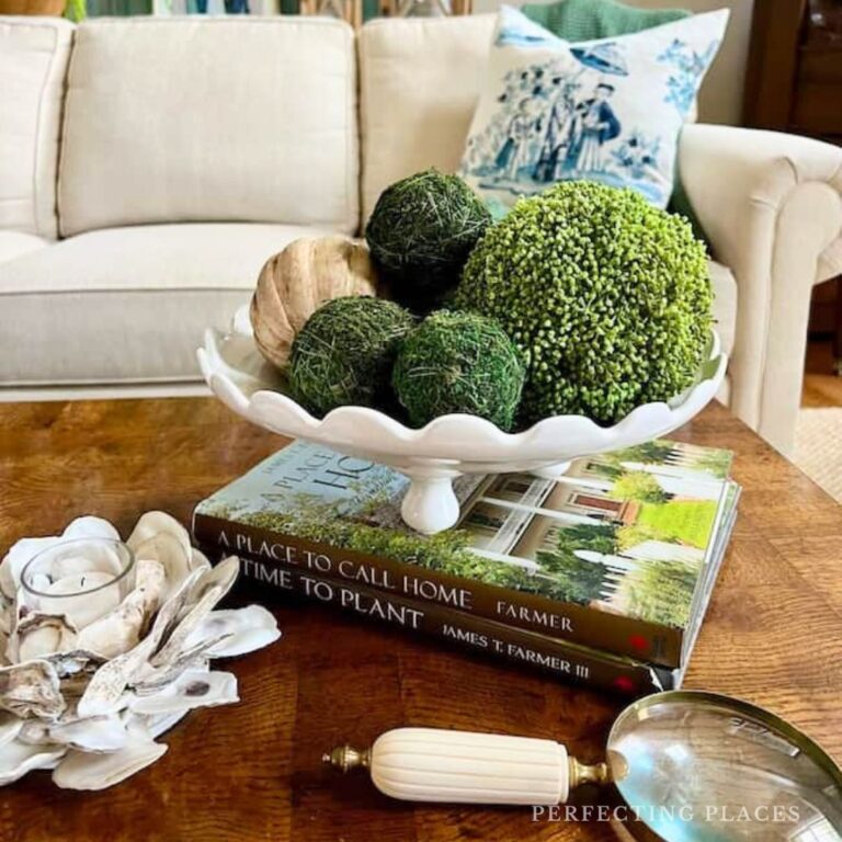A coffee table displays a white pedestal bowl styled to show how to decorate a footed bowl with decorative moss balls, two stacked books, a magnifying glass, and a shell centerpiece. A cream sofa with blue and green pillows is in the background.