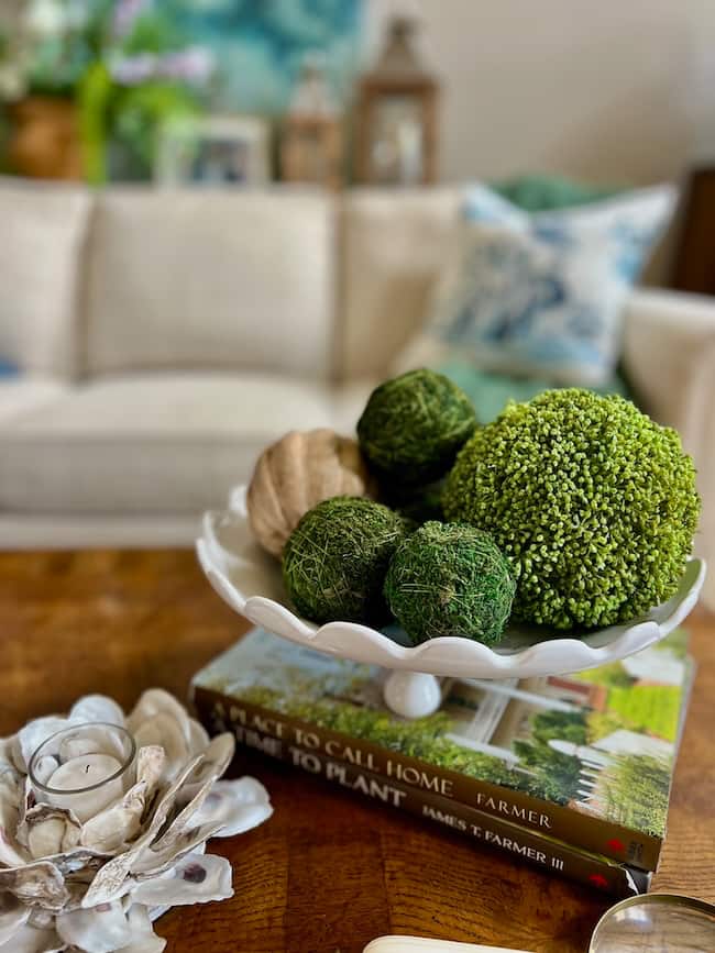 A white footed bowl is filled with green decorative moss balls and a tan accent, showing how to decorate a footed bowl on a wooden table, with books and a shell-shaped candleholder in front of a beige sofa with blue and white pillows.