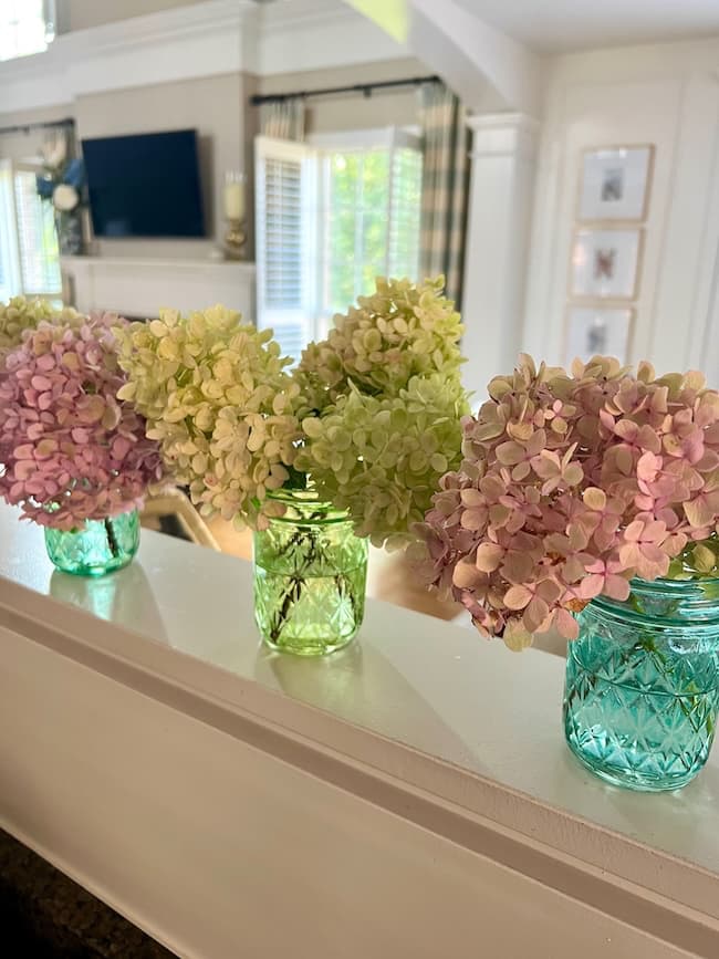 Three light green and pink hydrangea bouquets arranged in blue-green glass jars sit on a white ledge inside a bright, sunlit living room with neutral decor.